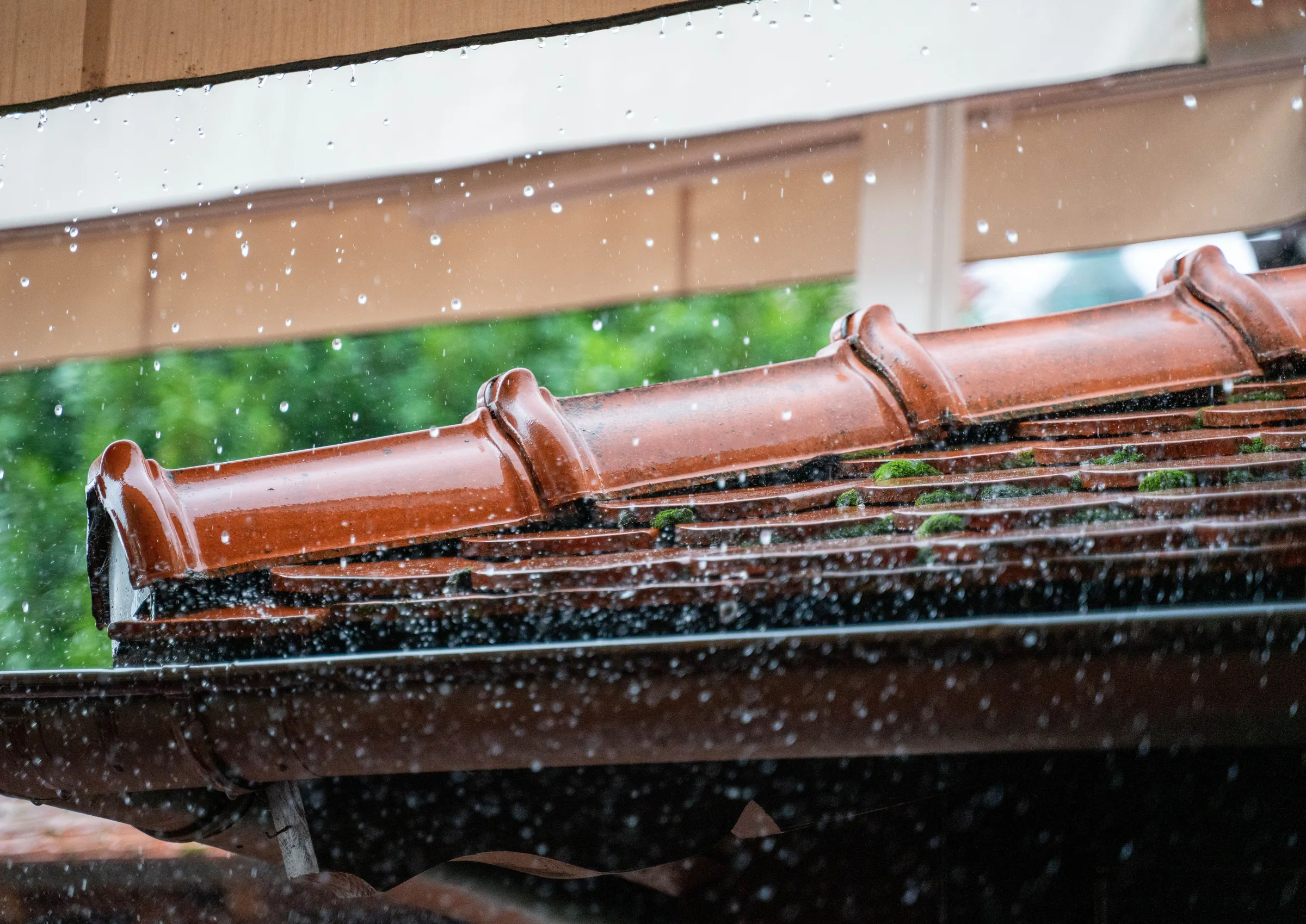 Rain falling on a gutter system with copper downspouts and green foliage visible in background.
