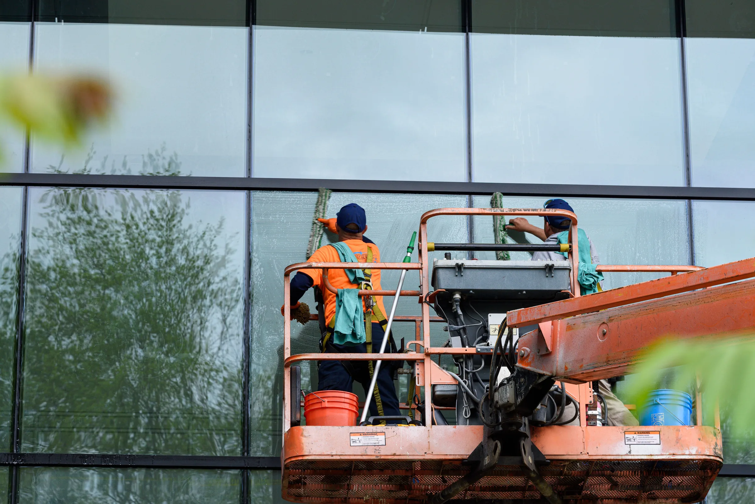 Two workers on an orange lift platform cleaning large glass building windows.